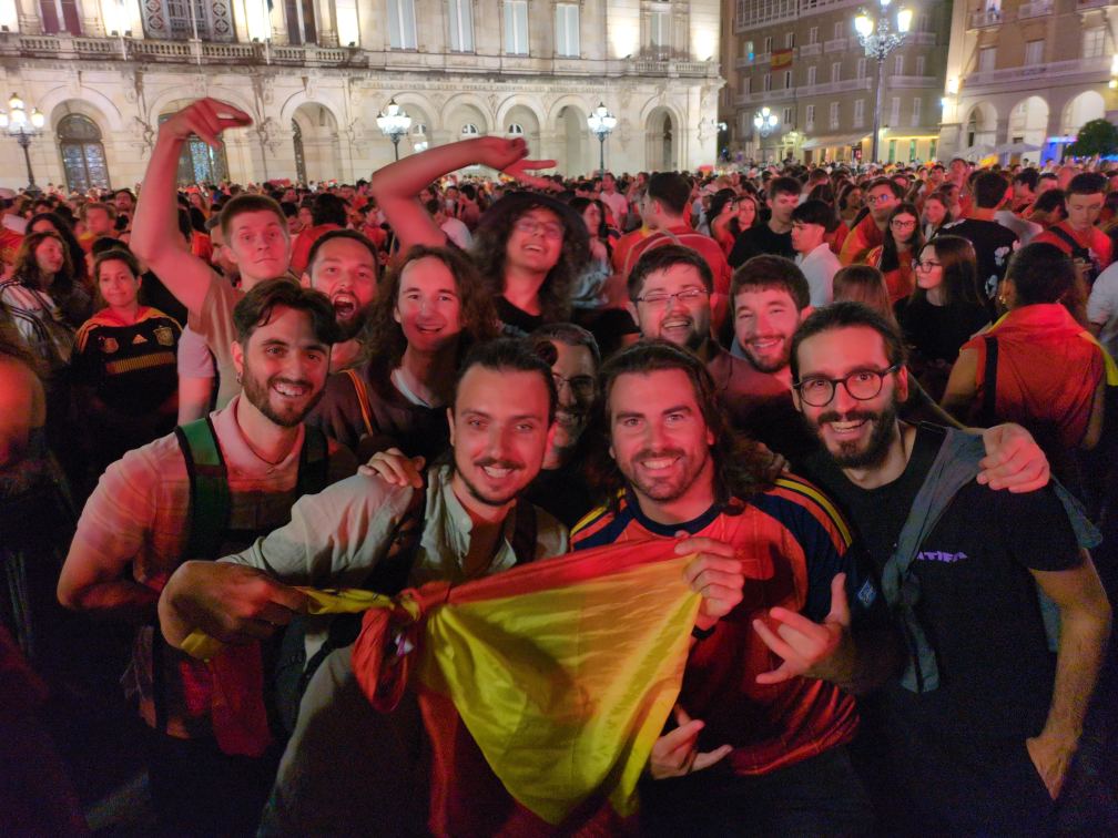 A group of around 10 GNOME foundation members (including me giving a very cheesy smile in the back c:) in middle of Plaza de María Pita, holding a Spanish flag celebrating Spain winning the world cup in a very large crowd.